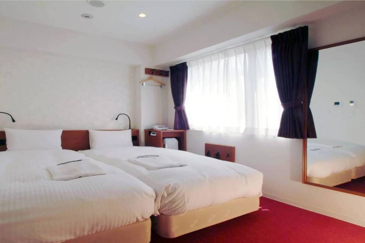 Ein Hotelzimmer mit zwei Betten, einem großen Spiegel, einem roten Teppich und einem Fenster mit Vorhängen.