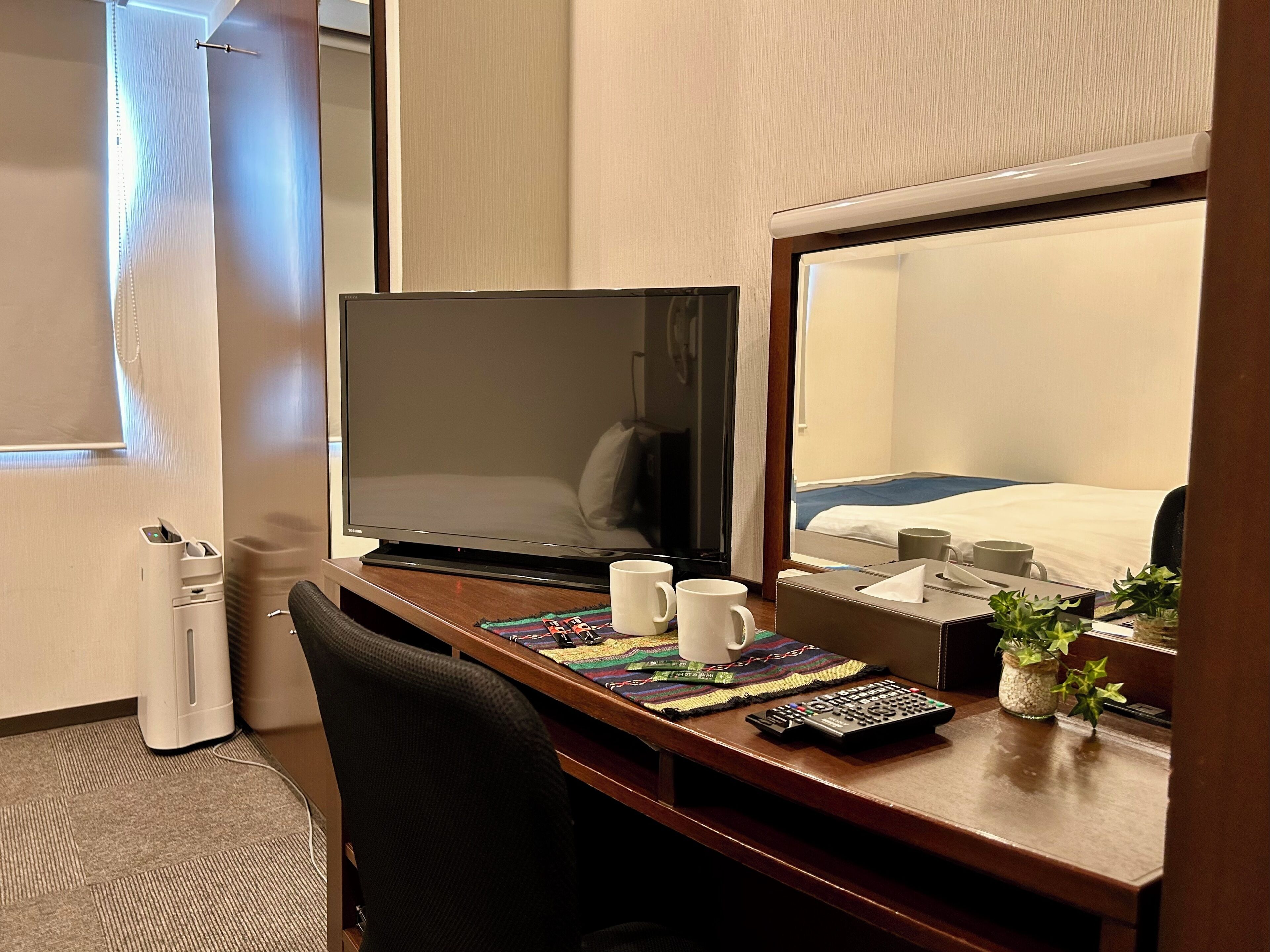 A hotel room with a desk, a laptop, a remote control, a tissue box, and a small plant.