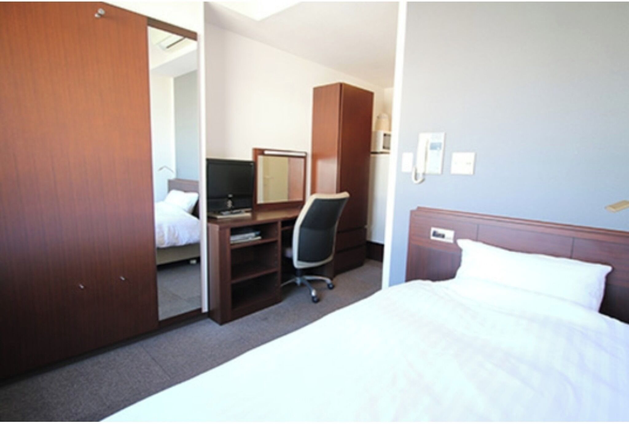 A hotel room with a bed, a desk with a chair, a wardrobe, and a mirror.
