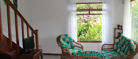 A room with a wooden staircase, two wicker chairs with patterned cushions, a small table, and a television on a stand.