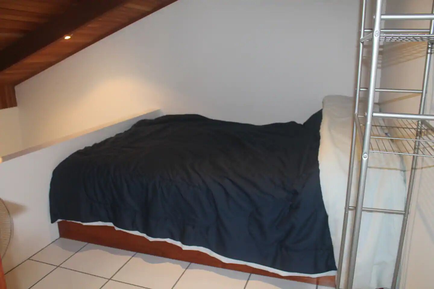 A single bed with a dark blue bedspread in a room with a sloped ceiling and a metal shelf unit to the right.