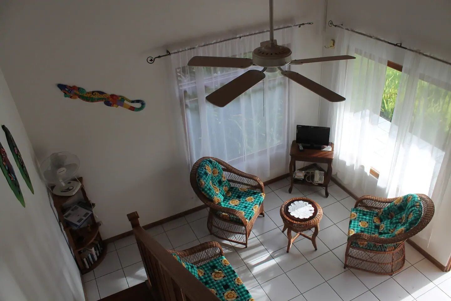 A room with a ceiling fan, two wicker chairs with floral cushions, a small round table, a television on a wooden stand, and a window with sheer curtains.