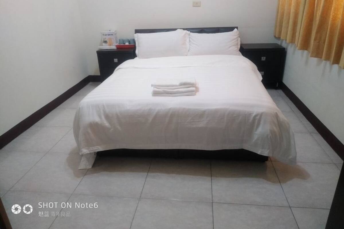 Cama bem arrumada com roupa de cama branca e uma toalha dobrada no rodapé. Um criado-mudo com abajur e uma pequena mesa ficam ao lado da cama.