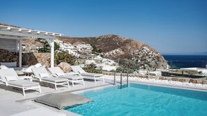 Zona de piscina con sillas de descanso blancas y una pérgola, con vistas a una colina con edificios blancos y al mar.