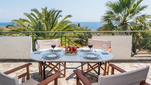 Un tavolo rotondo apparecchiato per due, con piatti, posate e bicchieri, affacciato su una vista mozzafiato del mare e delle palme.