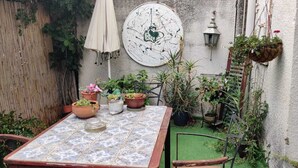 Um pequeno pátio com mesa de azulejos, vasos de plantas, um cachepot pendurado e um candeeiro fixado na parede.
