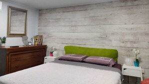 Quarto com parede de madeira, cabeceira verde, cama com colcha branca e roxa, cômoda de madeira, mesa de cabeceira com abajur e espelho.