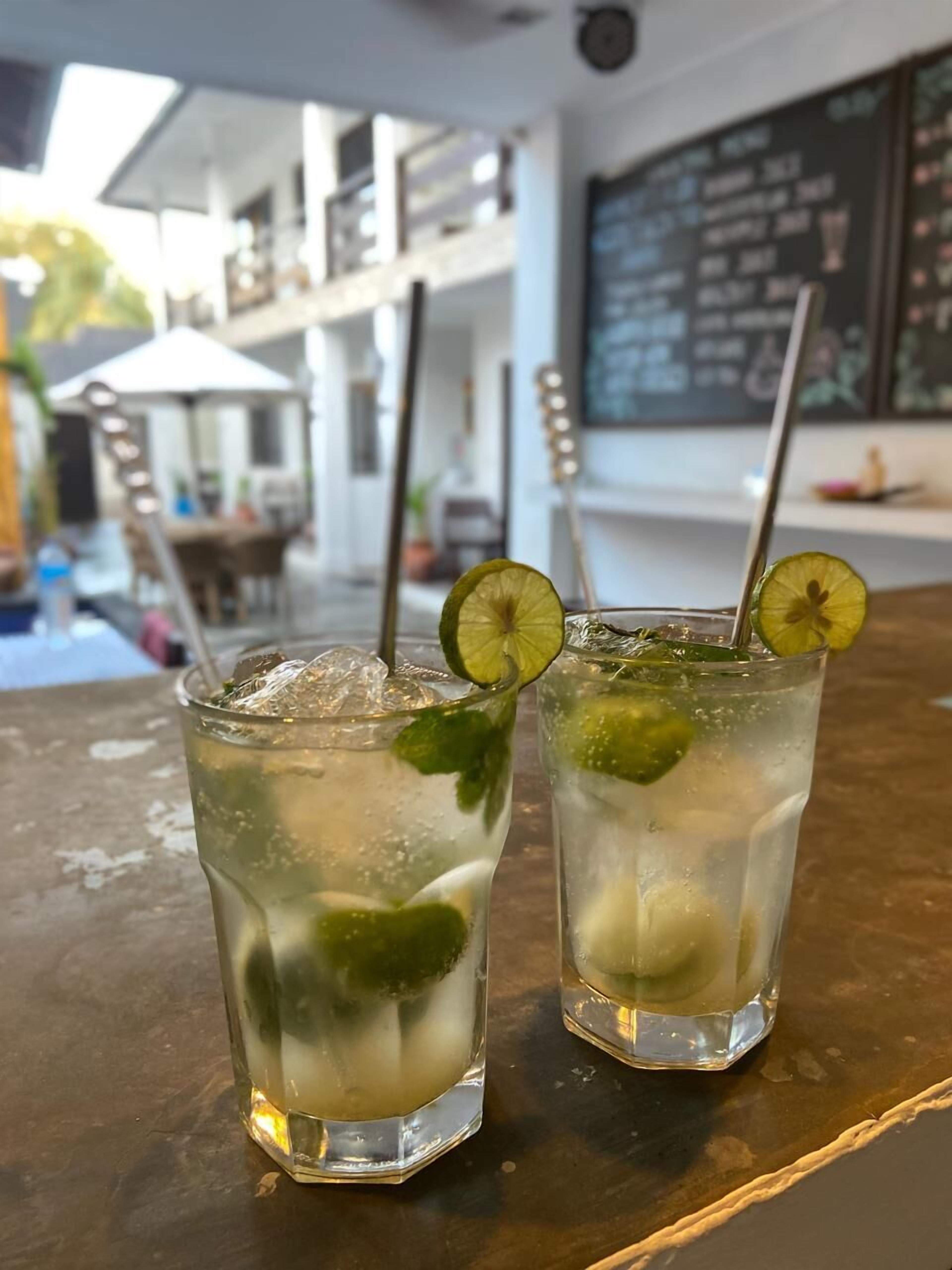 Deux verres contenant une boisson claire, agrémentée de rondelles de citron vert et de feuilles de menthe, sont posés sur une table en bois dans un restaurant. On aperçoit en arrière-plan un tableau noir servant de menu.