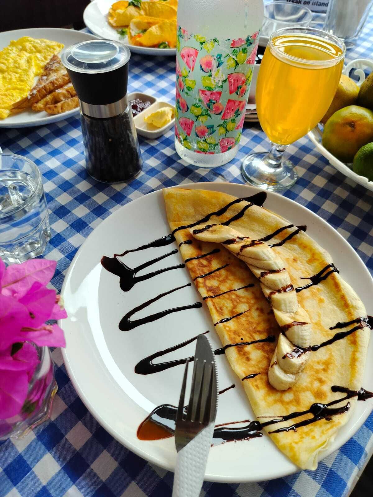 Une table mise pour le petit-déjeuner avec une crêpe, un verre de jus d’orange et une bouteille d’eau, le tout posé sur une nappe à carreaux.