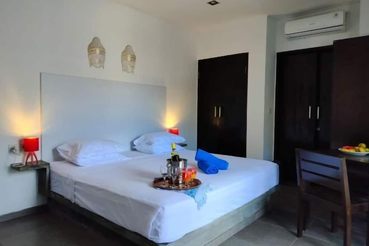 A hotel room with a bed, bedside lamps, a minibar, a desk with a chair, and a wardrobe.
