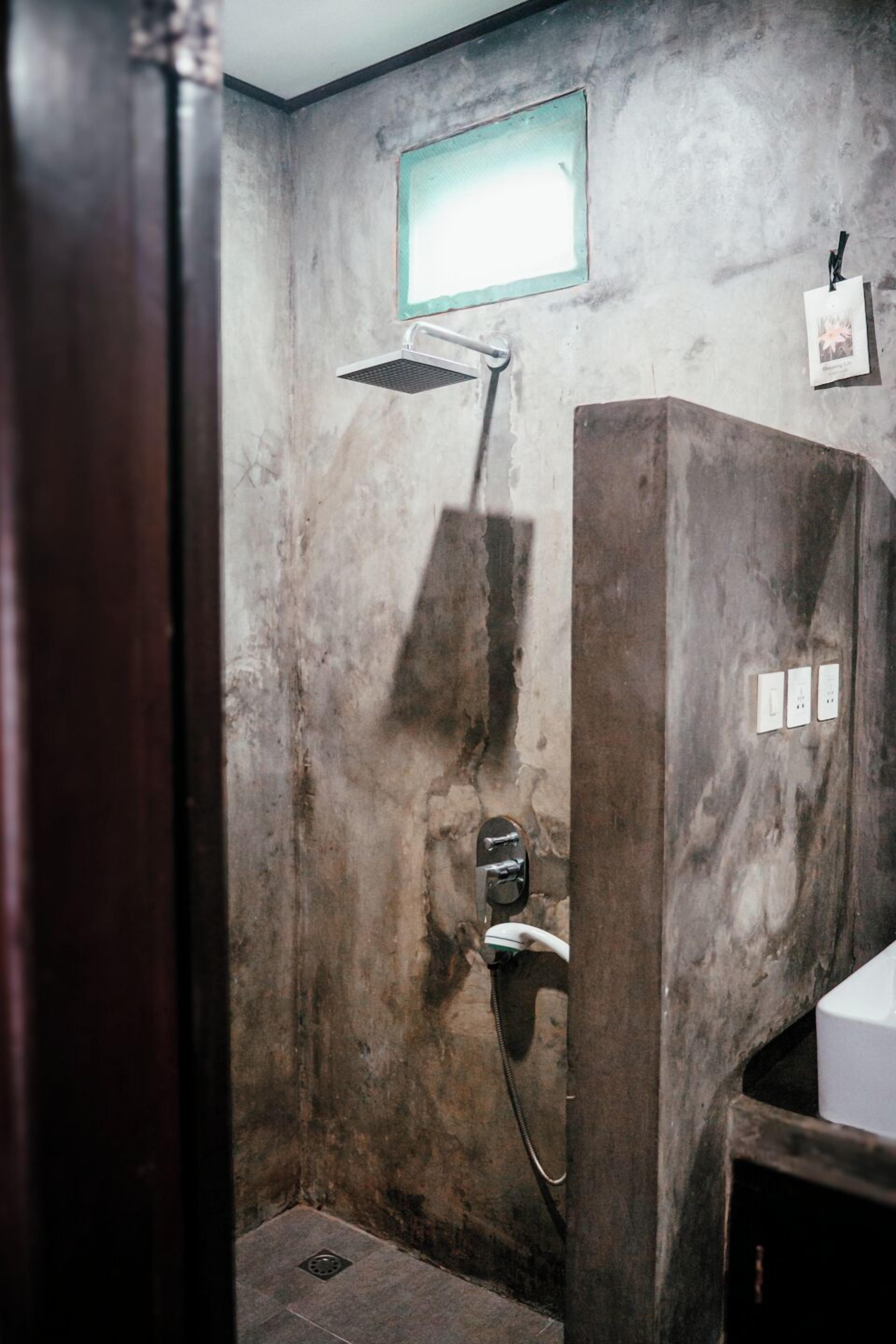 Um banheiro com parede de concreto, chuveiro e uma pequena janela.