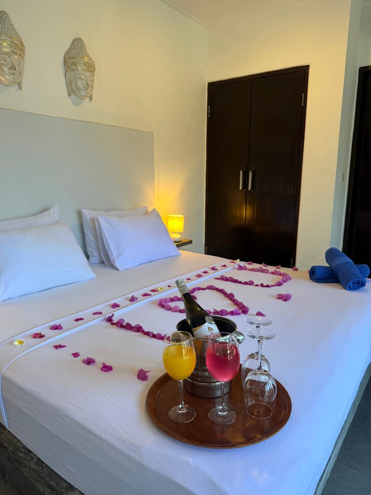 Quarto de hotel com cama, mesa de cabeceira com abajur, minibar e bandeja com champanhe, duas taças e um suco de laranja.