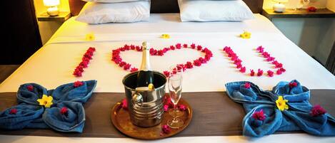 A hotel room with a bed, pillows, a bedside table with a lamp, a bottle of champagne, a glass, and towels arranged on the bed.