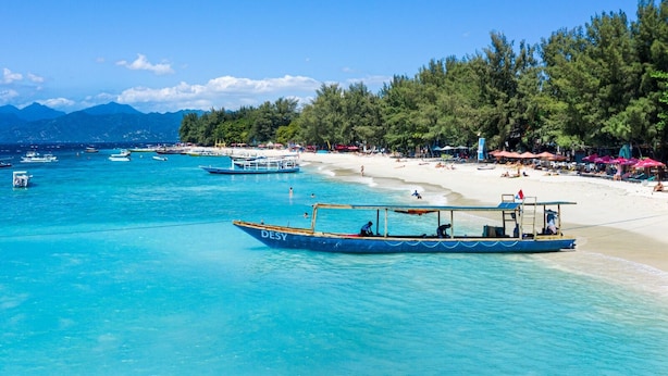 Ein Strand mit klarem türkisblauem Wasser, Booten und einem sandigen Ufer, das von Bäumen und Sonnenschirmen gesäumt wird.