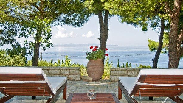 A terrace with two wooden lounge chairs and a table, overlooking a body of water with a stone wall and a large planter with red flowers.