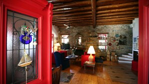 Una sala de estar acogedora con pared de piedra, vigas de madera en el techo y un ventanal con marco rojo y objetos decorativos.
