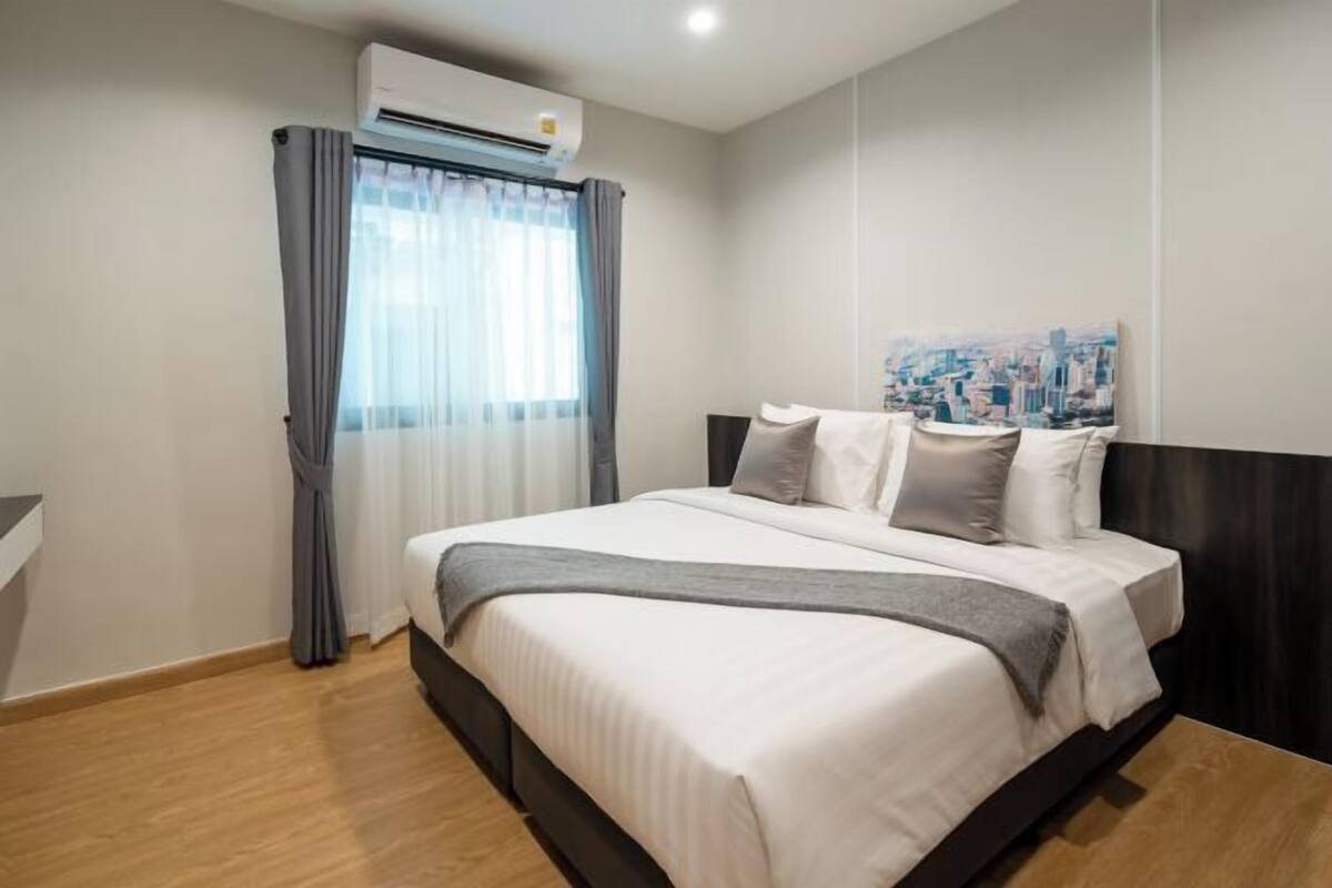 A hotel room with a bed, curtains, an air conditioning unit, and a cityscape painting on the wall.