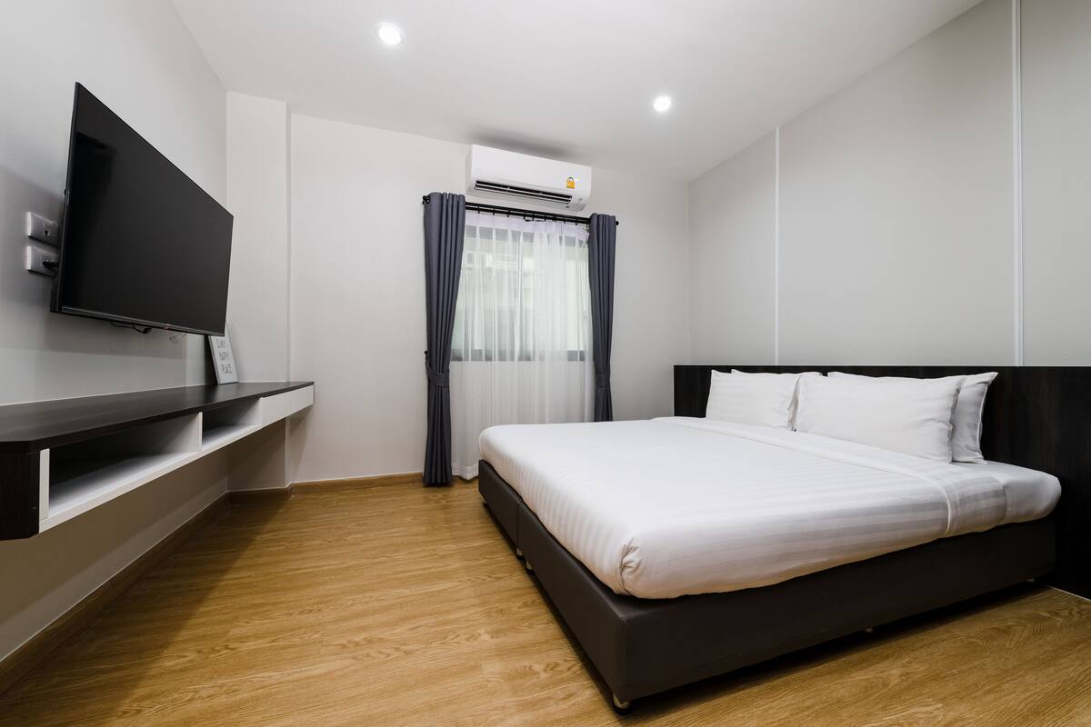 A modern hotel room with a flat-screen TV, a bed with white bedding, a dark headboard, and a wooden floor.