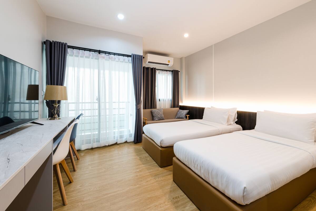 A hotel room with two beds, a desk, a chair, a TV, and an air conditioning unit.