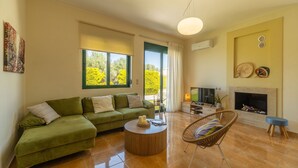 A living room with a green sofa, a round coffee table, a wicker chair, a fireplace, and a television. - Holiday villa with private pool (Ξηροκάμπι)