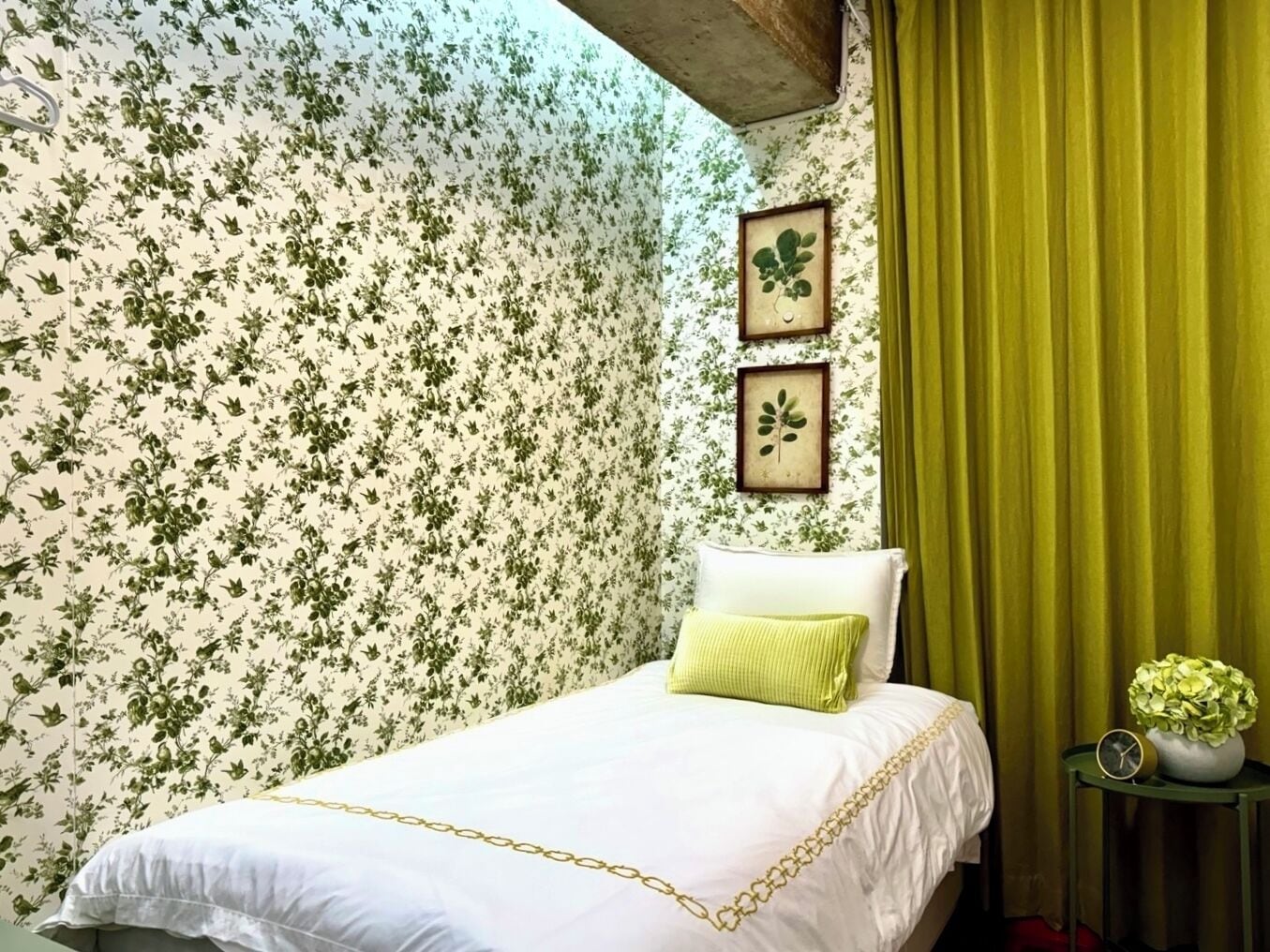 Une chambre avec un papier peint à motifs floraux, un lit recouvert d’une courtepointe blanche et dorée, un coussin vert et une petite table sur laquelle se trouvent une horloge et une plante.