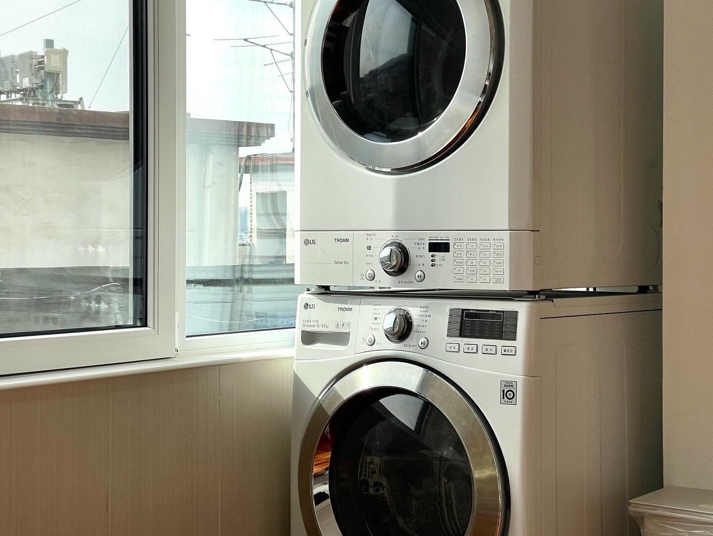 Een wasruimte met twee gestapelde wasmachines en een droger, een raam met uitzicht op gebouwen en een houten vloer.
