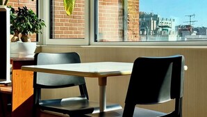Ein Essbereich mit Tisch und zwei Stühlen, einer Topfpflanze und Blick auf ein Backsteingebäude durch ein Fenster.