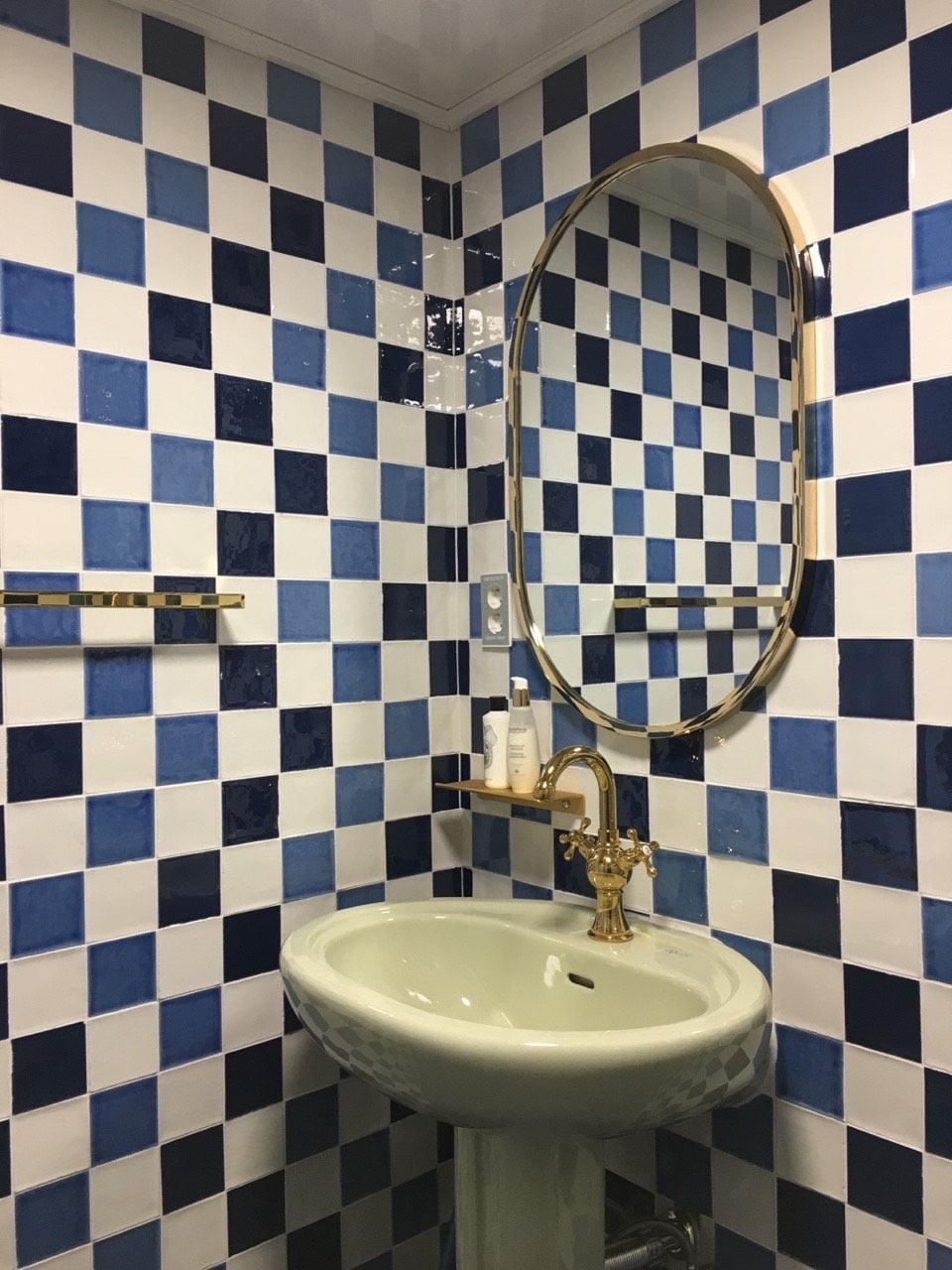 Ein Badezimmer mit Wandverkleidung aus blau-weiß karierten Fliesen, einem Standwaschbecken, einem goldenen Wasserhahn und einem runden Spiegel.
