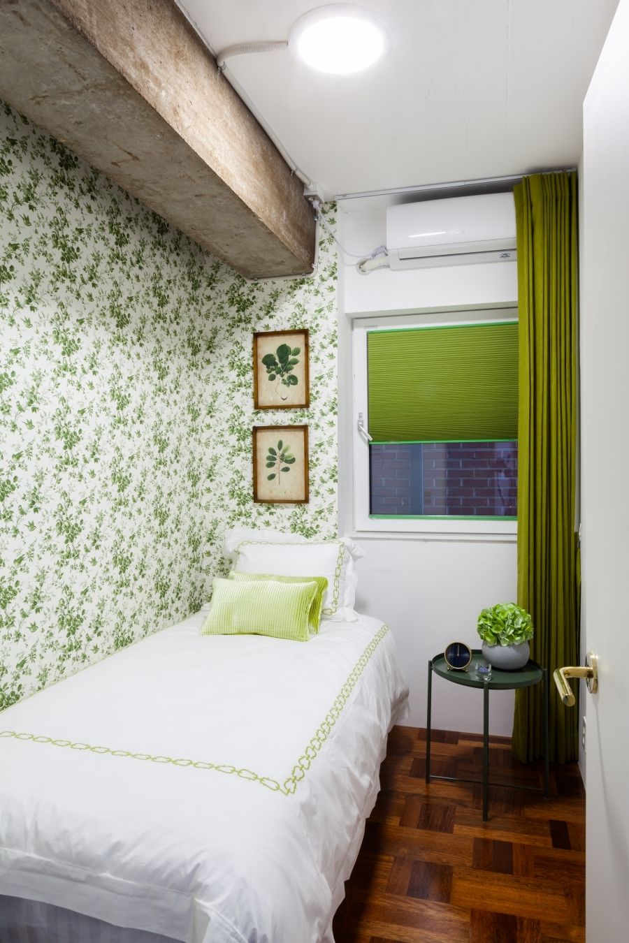 Un dormitorio con cama, una mesita con una planta, una ventana con cortinas verdes y un aire acondicionado instalado en la pared.