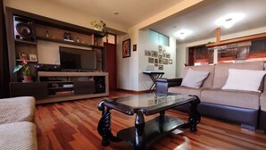 Sala de estar com mesa de centro de vidro, sofá, televisão e área de cozinha ao fundo.