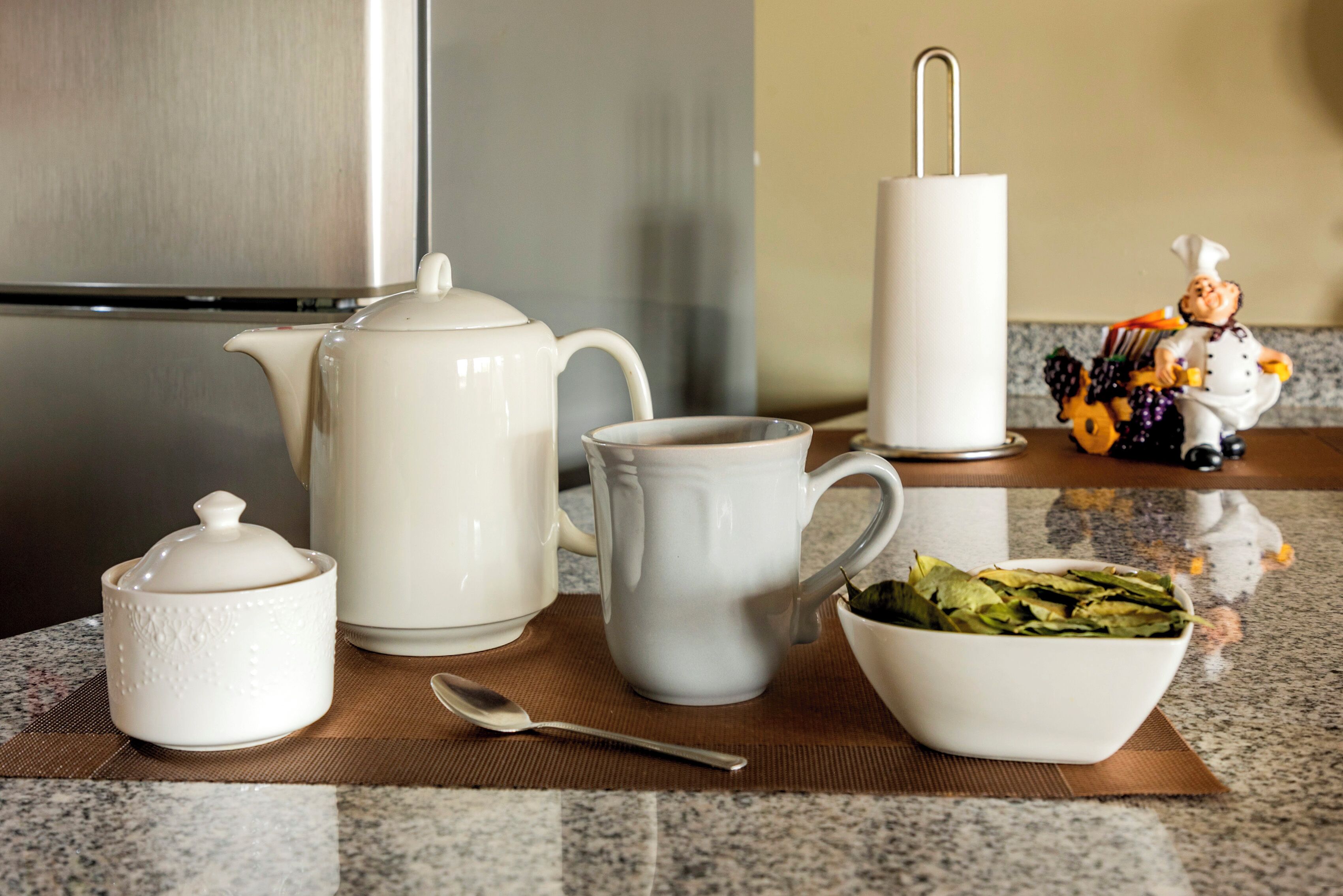 Una encimera de cocina con una tetera blanca, una jarra blanca, un cuenco blanco con verduras y un azucarero, además de una cuchara.