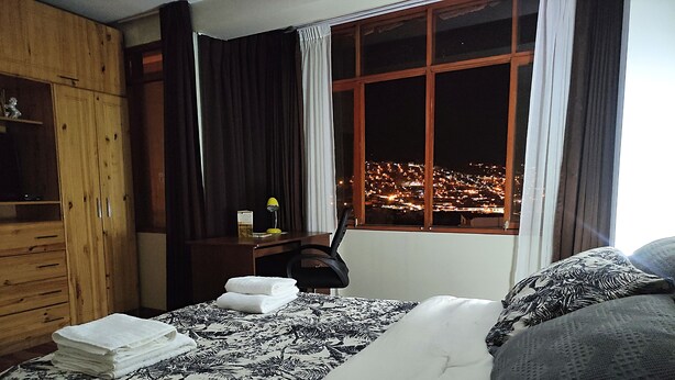 Quarto de hotel com cama, escrivaninha, cadeira, guarda-roupa e janela com vista para a cidade à noite.