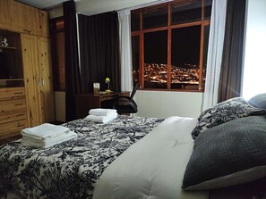 A hotel room with a bed, a desk, a chair, a wardrobe, and a window with a city view at night. - El Balcón del Sol (Cusco)