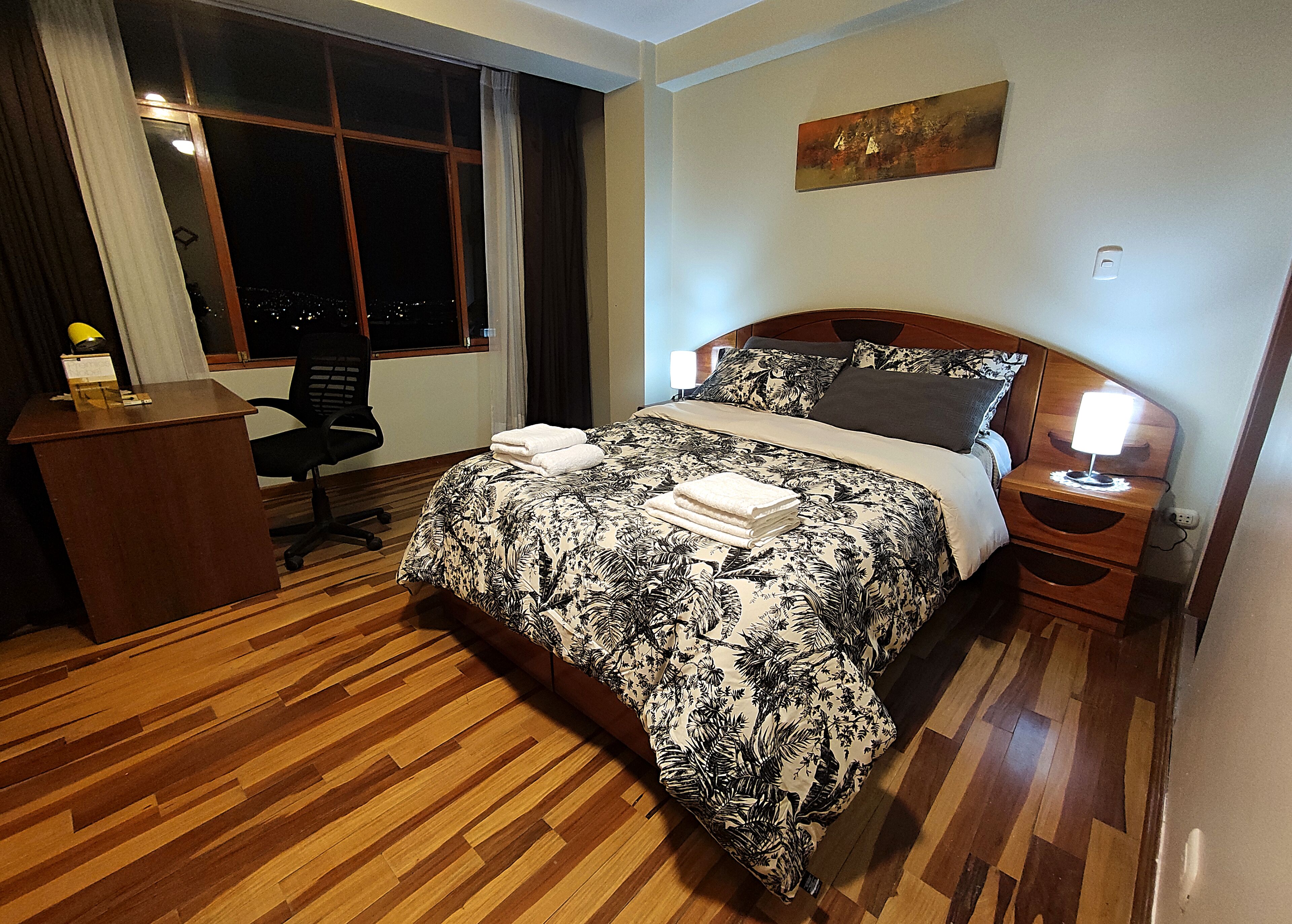 A hotel room with a wooden floor, a bed with a floral bedspread, a desk with a lamp, and a nightstand with a lamp.