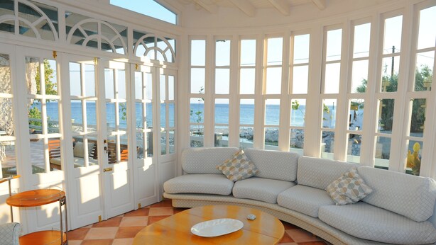 Una stanza luminosa con pavimento a scacchi, un divano con cuscini decorati e vista sul mare attraverso ampie finestre.