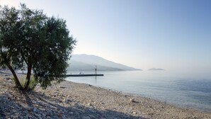 Una spiaggia di ciottoli con un faro all'orizzonte, acqua calma e una catena montuosa.
