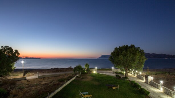 A nighttime view of a coastal property with a well-lit pathway, a grassy lawn, and a view of the sea and distant mountains. - Waterfront apartment 4 (Κίσσαμος)