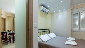 A bedroom with a bed, an air conditioning unit, a dining area with chairs and a table, and a window with blinds. - Waterfront apartment 4 (Κίσσαμος)