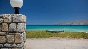 A stone wall with a spherical light fixture, a dirt path, and a small boat on a turquoise sea with distant mountains. - Waterfront apartment 4 (Κίσσαμος)