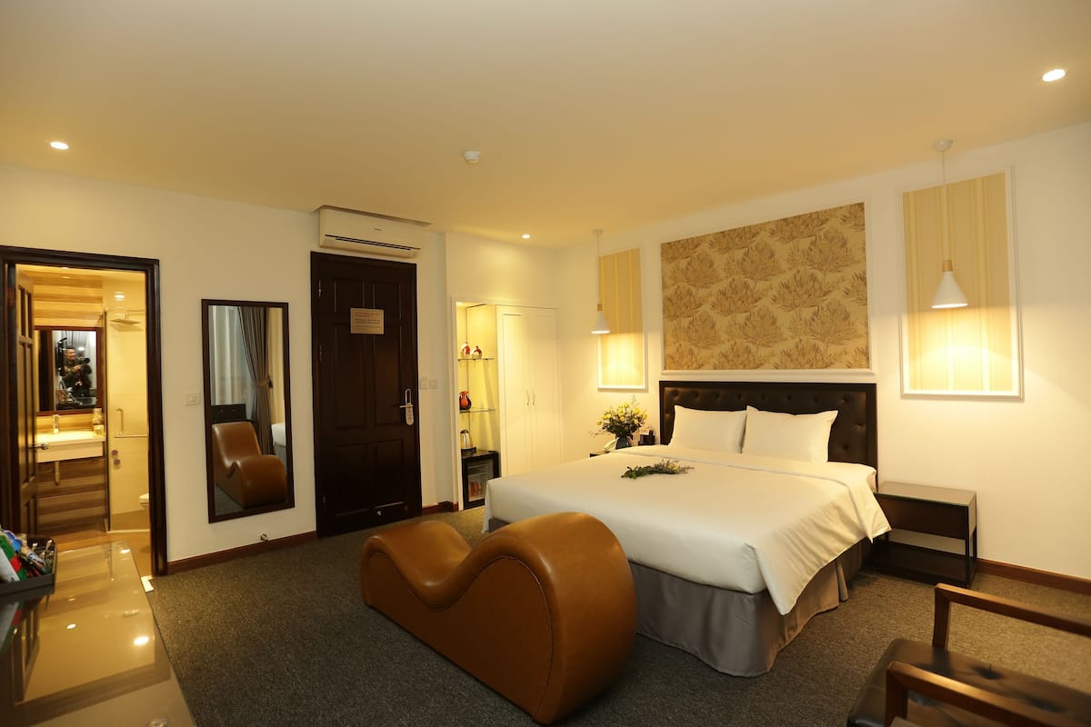 A hotel room with a bed, a seating area, a mirror, a nightstand, and a wall-mounted lamp.