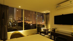 Habitación de hotel con un ventanal grande que ofrece vistas de la ciudad de noche, un televisor de pantalla plana montado en la pared y una zona de comedor con mesa y sillas.