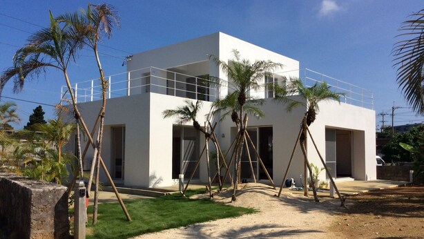 Un edificio moderno de dos pisos con fachada blanca, rodeado de palmeras y un área verde.