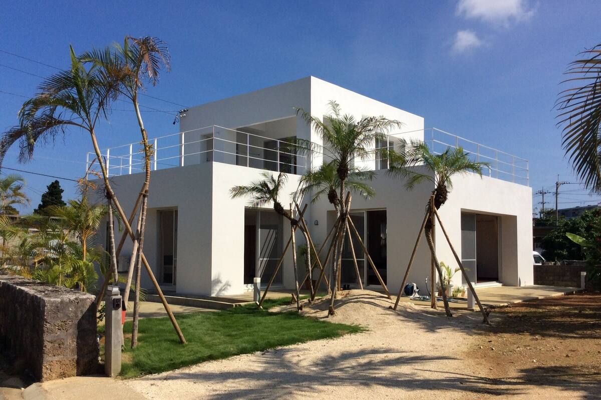 A modern two-story building with a white exterior, surrounded by palm trees and a grassy area.