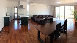 Una cocina moderna con una isla central, un comedor con mesa y sillas de madera, un conjunto de sofás y ventanas amplias que dejan entrar luz natural.