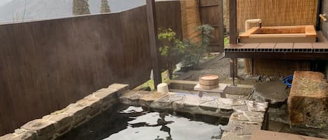 A hot spring area with stone steps leading into a pool, surrounded by wooden structures and a view of mountains.