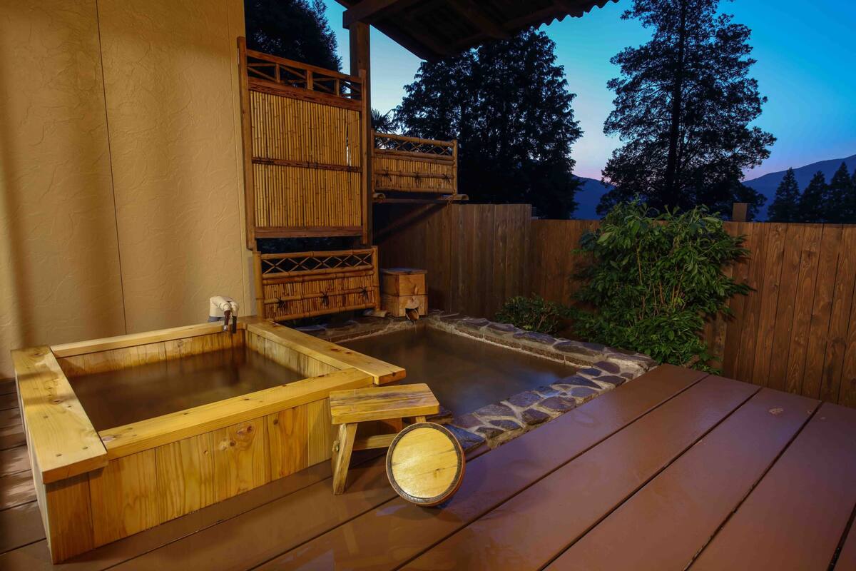 A wooden hot tub with a stone surround, a wooden deck, and a wooden fence with a bamboo gate.