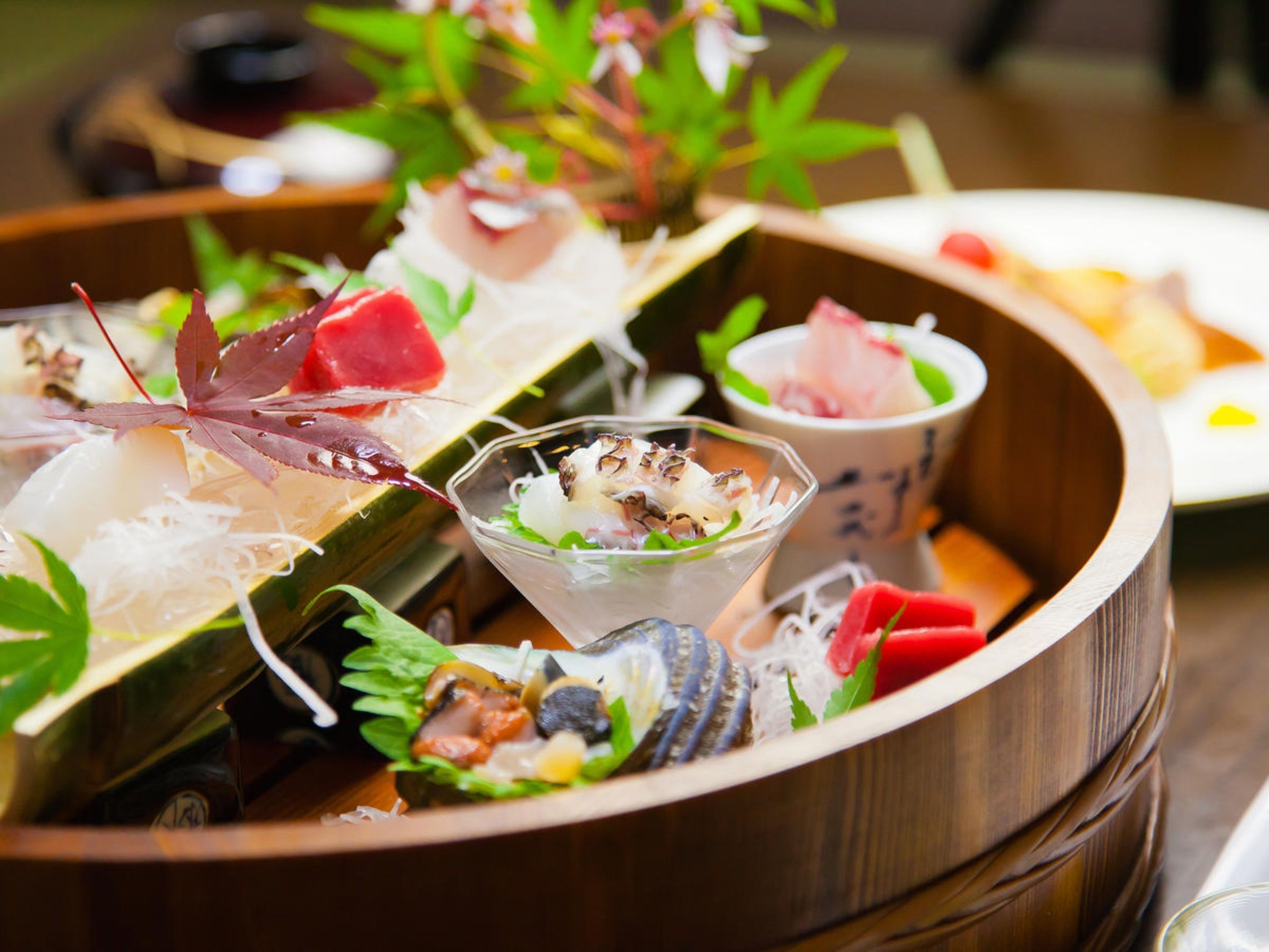 A wooden serving dish with various sushi and sashimi dishes, including a bowl with a decorative leaf.