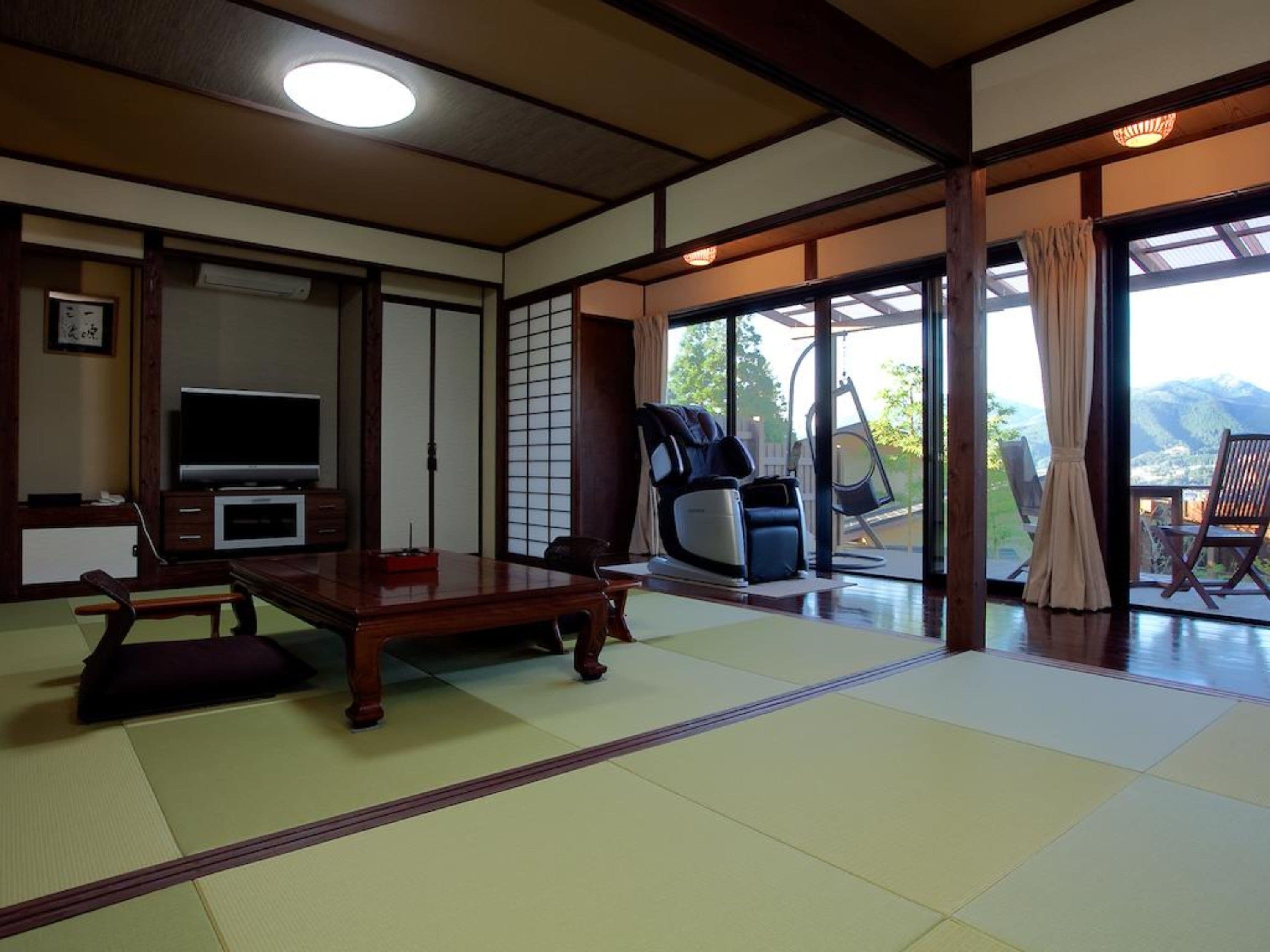 Una habitación japonesa tradicional con suelo de tatami, una mesa baja de tatami, televisión, sillón de masaje y puerta corredera de cristal que da a un balcón con vistas a la montaña.