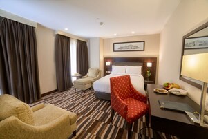Habitación de hotel con cama, escritorio con lámpara, silla y mesa de comedor con frutas y un plato.