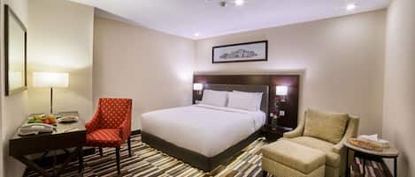 A hotel room with a large bed, two armchairs, a desk with a lamp, and a framed picture on the wall.